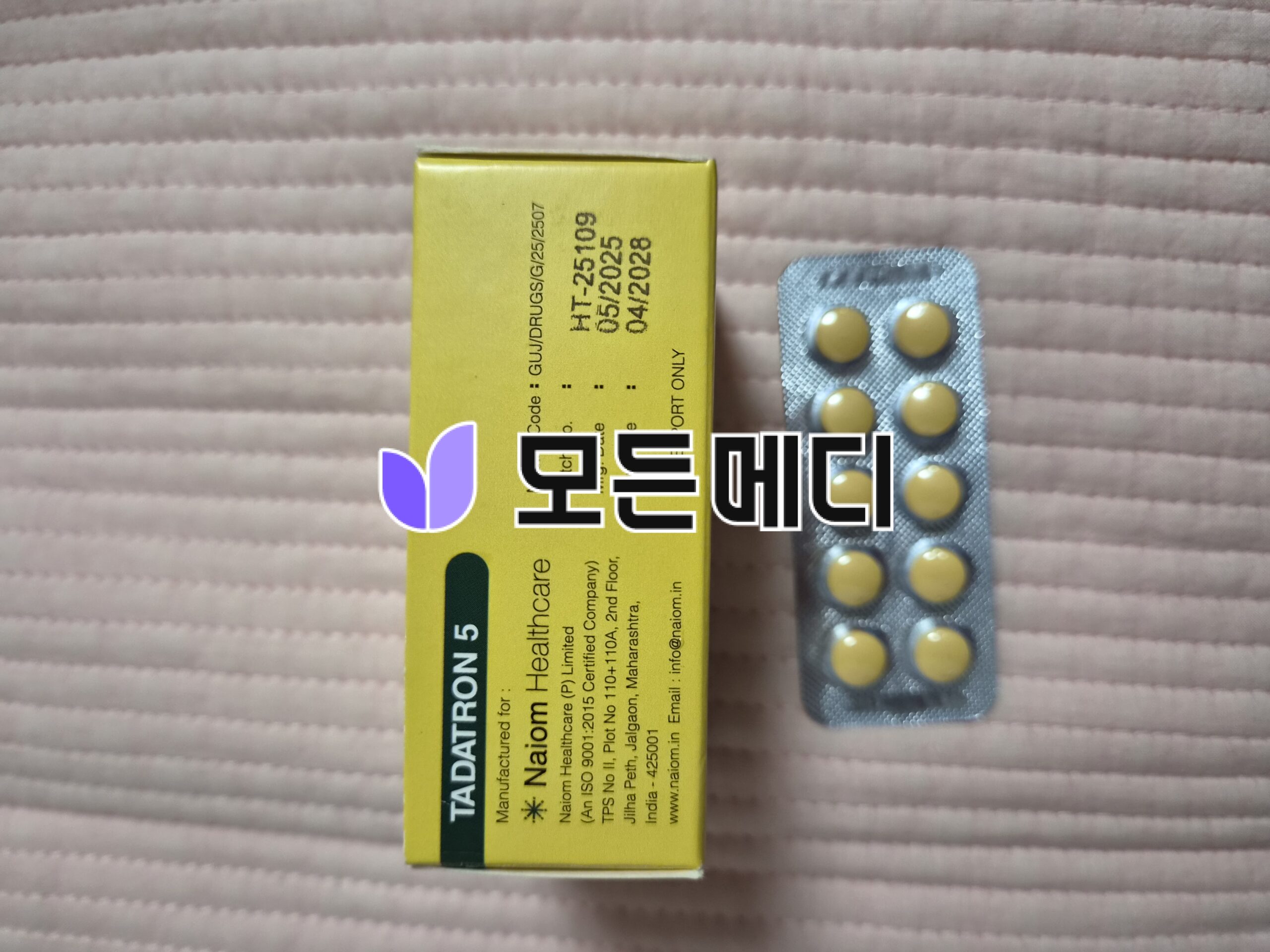 Yellow medicine box beside a blister pack of round yellow tablets on a pink quilted surface, showing pharmaceutical packaging.