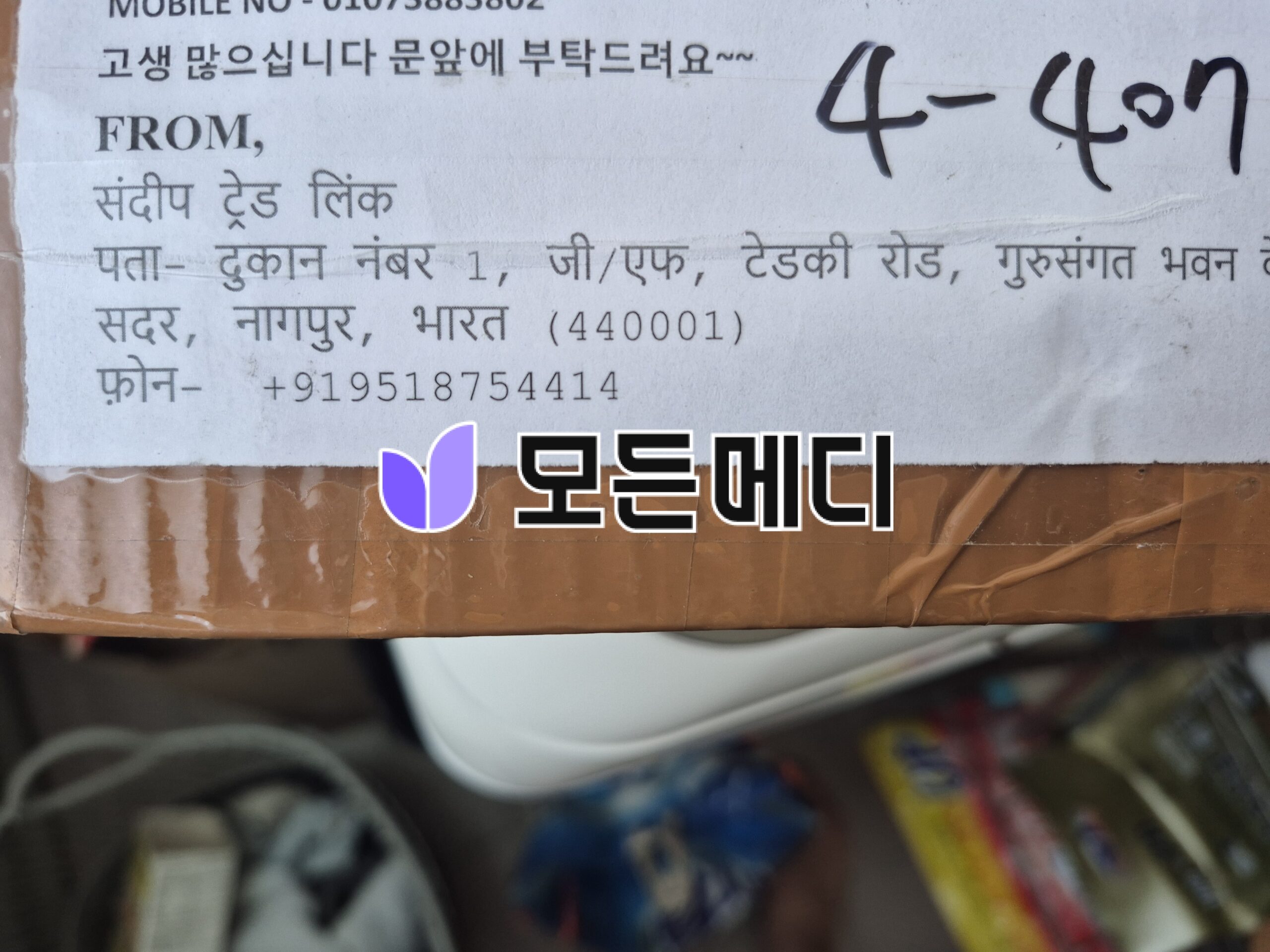 Close-up of a cardboard package label with printed and handwritten text including FROM, address details in Hindi, and a purple logo overlaid near the bottom right.