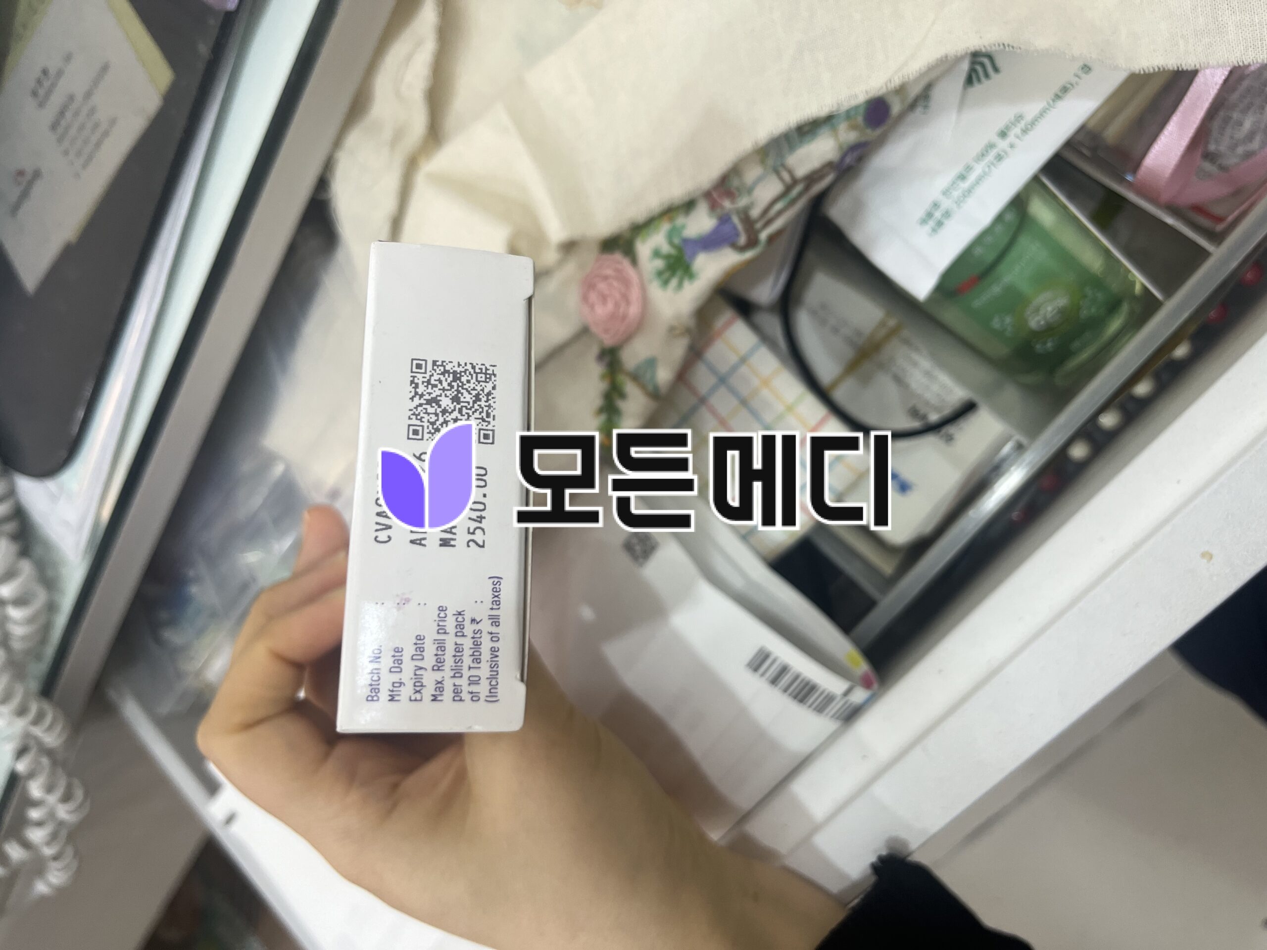 Person holding a white medicine box with a QR code and purple logo, in a pharmacy aisle near shelves and other boxes.