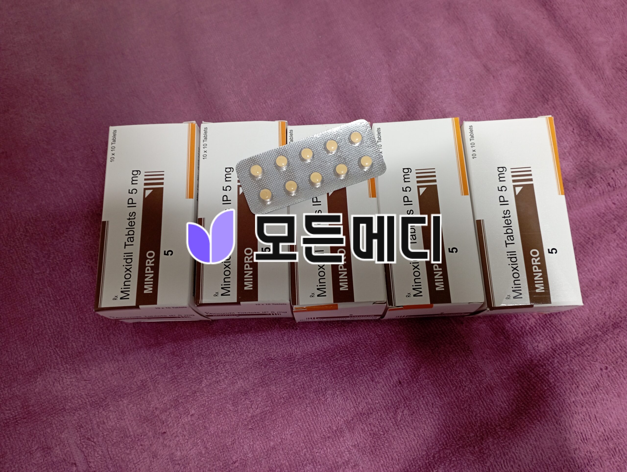 IMG_20260417_214458 | 모든메디 Row of Minoxidil 5 mg tablet boxes arranged in a row with a blister pack resting on top, on a purple fabric background.