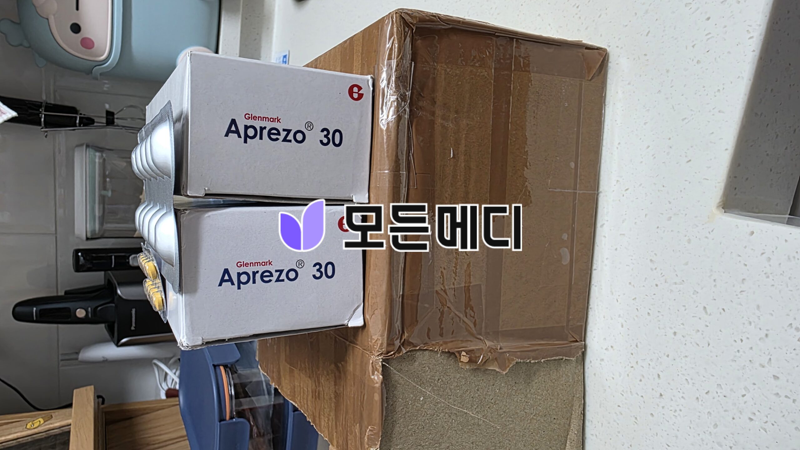 Two Glenmark Apreso 30 boxes stacked on a brown cardboard box in a cluttered room shelf area.