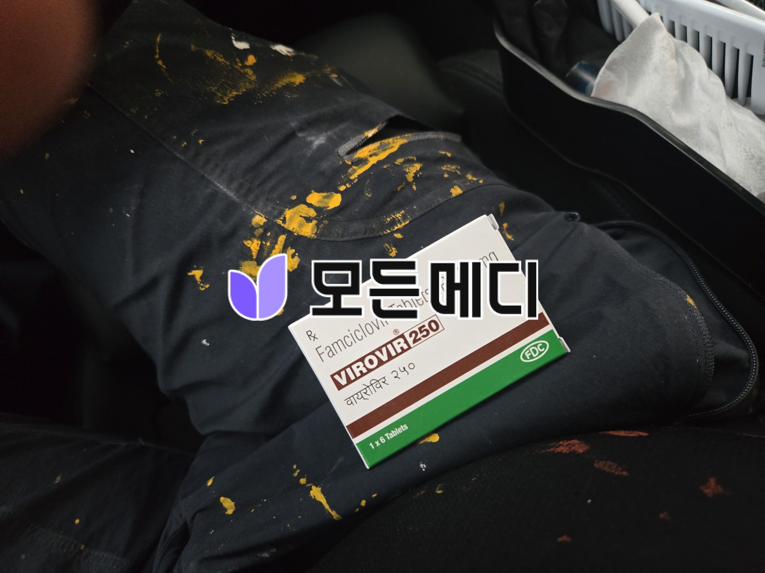 Close-up of black fabric with yellow paint splatters and a boxed medicine labeled Famciclovir 250 mg with a purple logo nearby.