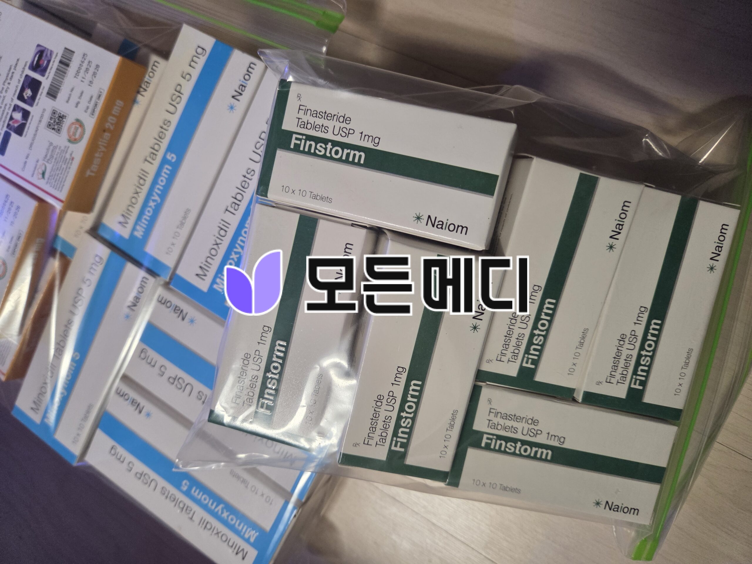 Assorted medicine boxes in a plastic bag on a wooden surface, including Finastorm Finasteride 1 mg tablets.