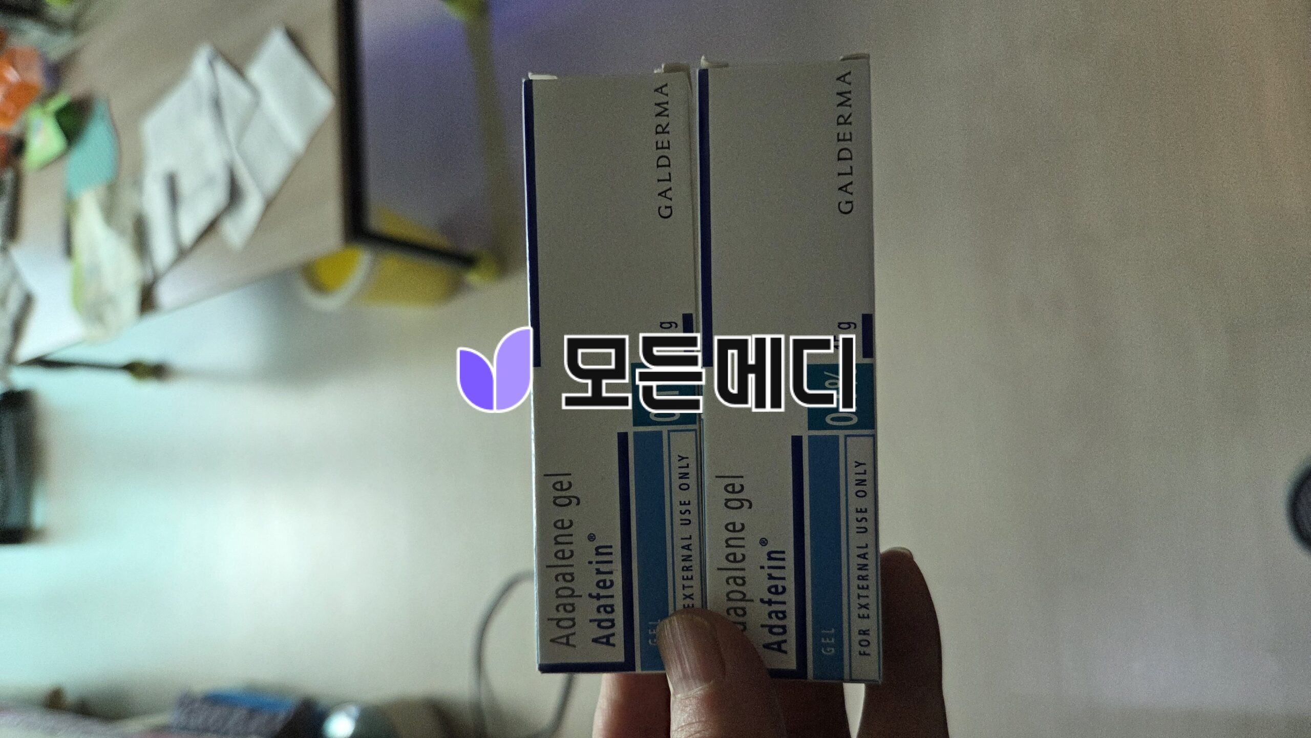 Two boxes of Adapalene gel acne treatment held in a hand, labels read 'Adapalene gel' vertically on white boxes with blue accents (external use).