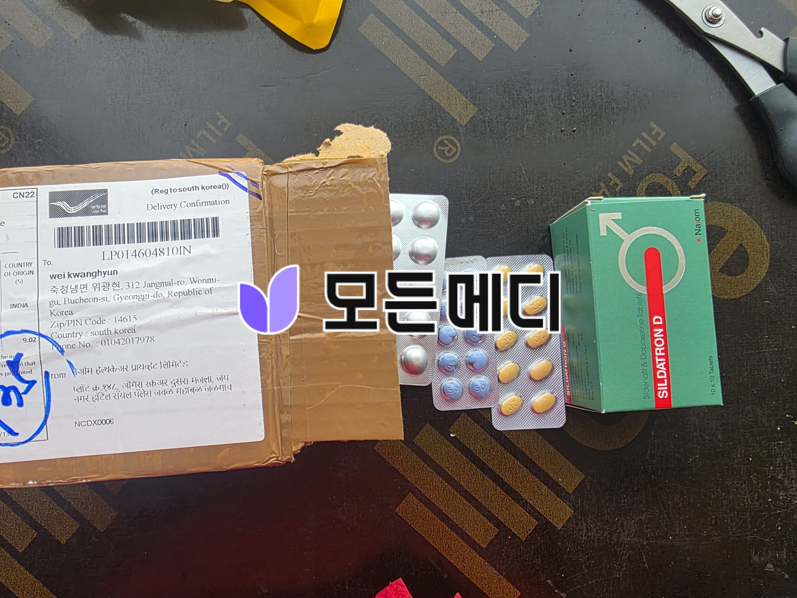 Top-down view of a cardboard package with a delivery label, blister packs of pills, and a green medicine box on a dark surface