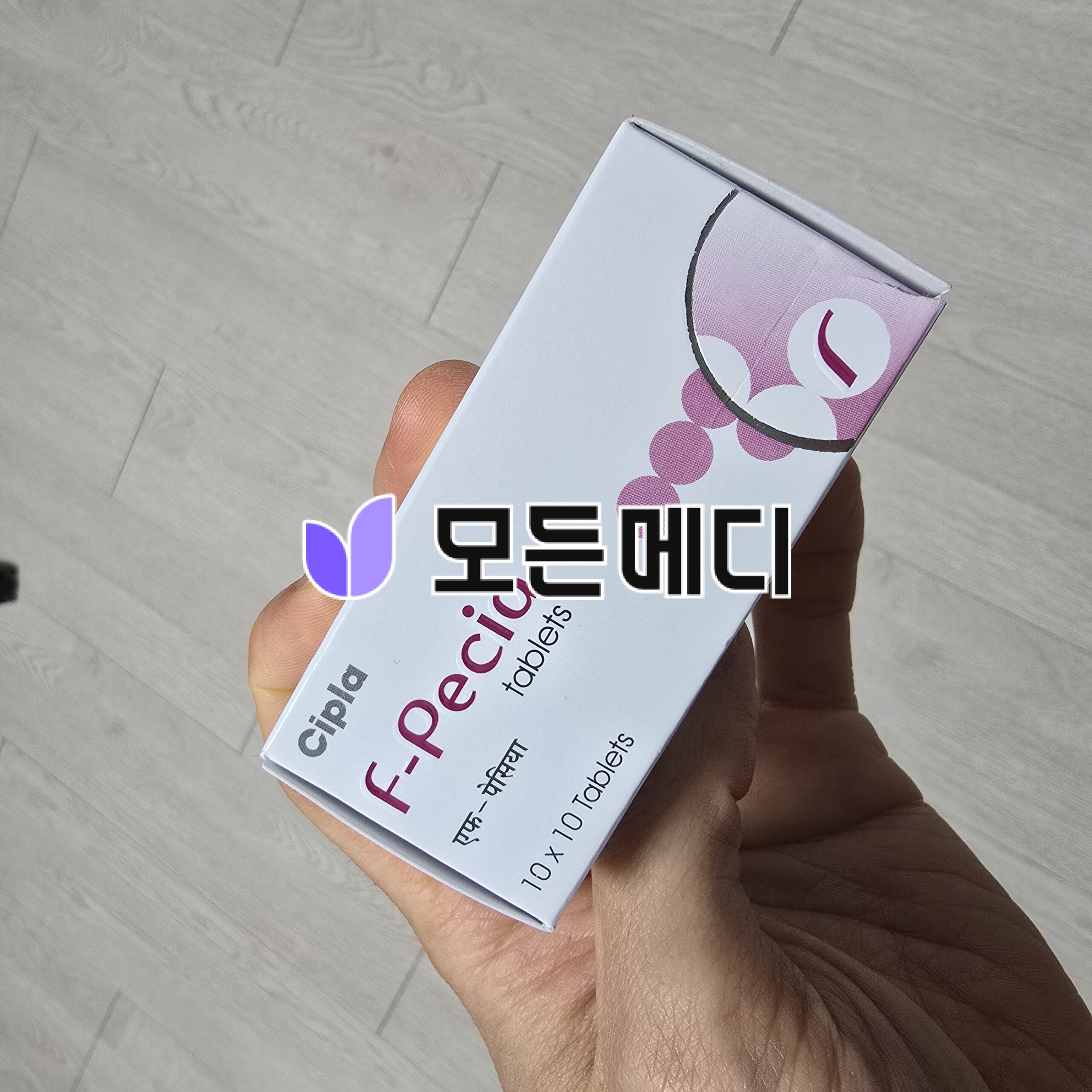 Hand holding a light blue box with pink circular graphic, against a tiled floor background.
