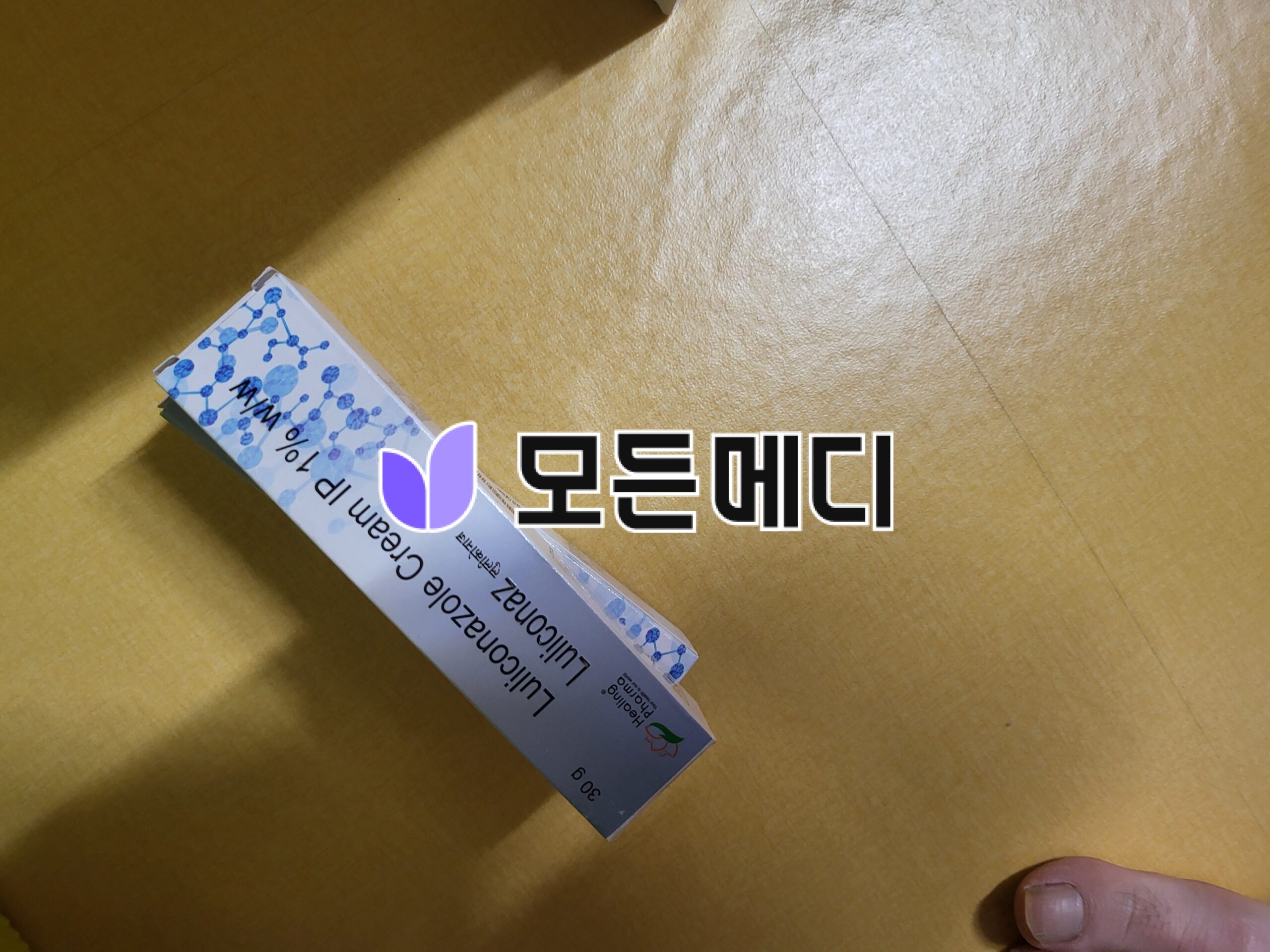 Box of a dietary supplement with blue molecular graphics and a purple leaf logo lying diagonally on a wooden floor; a fingertip appears in the bottom right.